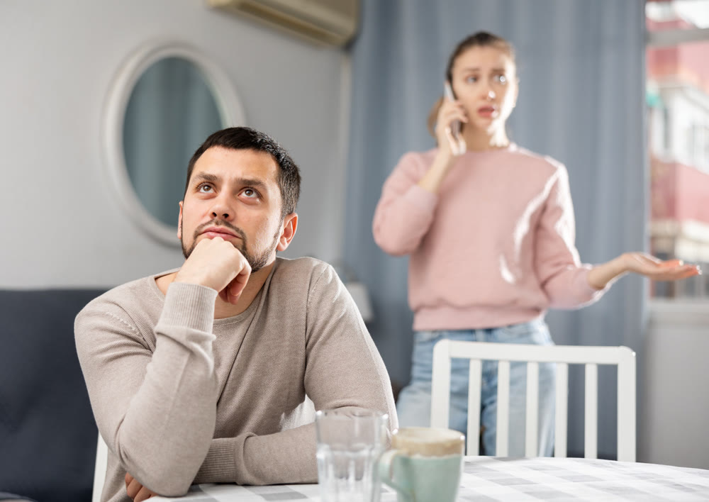 Sad man sitting at table at home, wife talking on phone
