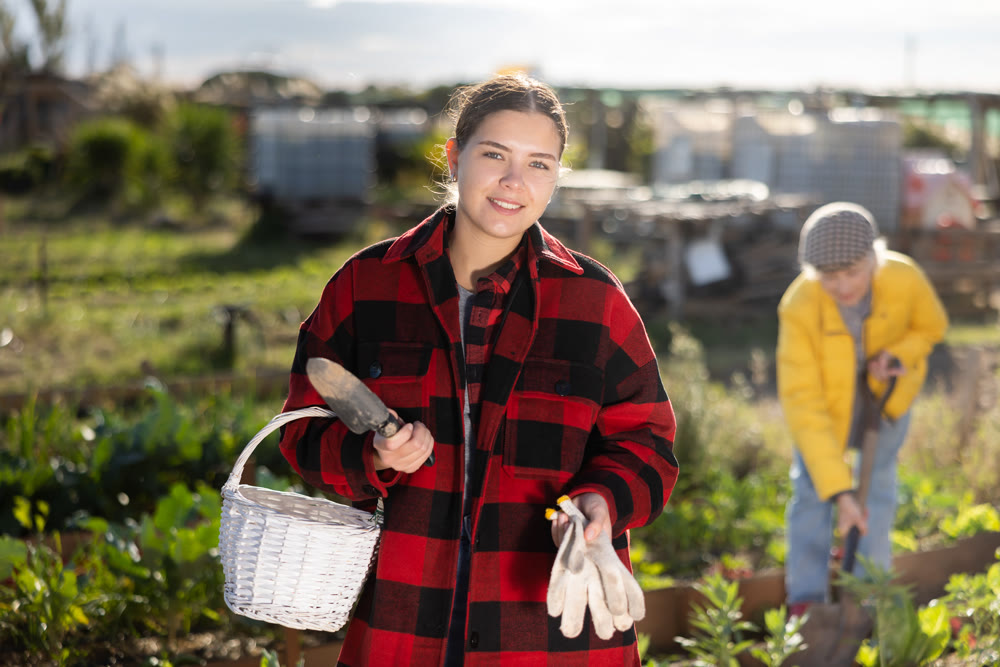Smiling young female farmer posing with basket and garden instruments during season of sowing on sunny day of autumn