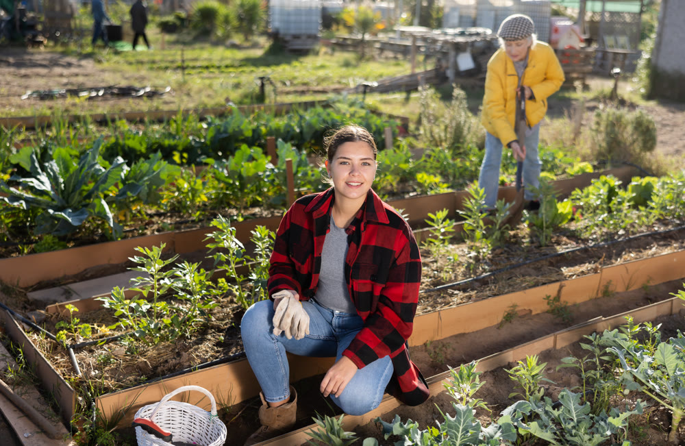 Smiling young female farmer posing with basket and garden instruments during season of sowing on sunny day of autumn