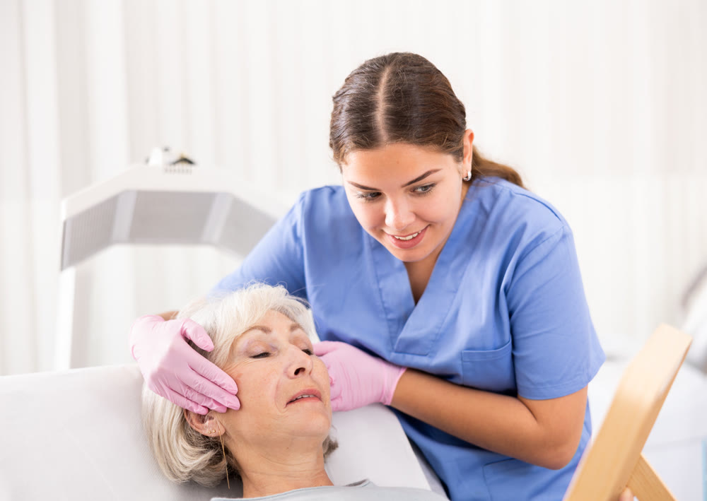 Satisfied senior woman looking at mirror after professional cosmetology treatment with beautician in modern beauty salon indoors