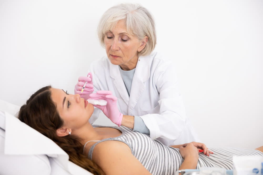 Elderly female doctor cosmetologist making rejuvenating injections procedure for tightening and smoothing wrinkles on face skin of young woman in beauty salon
