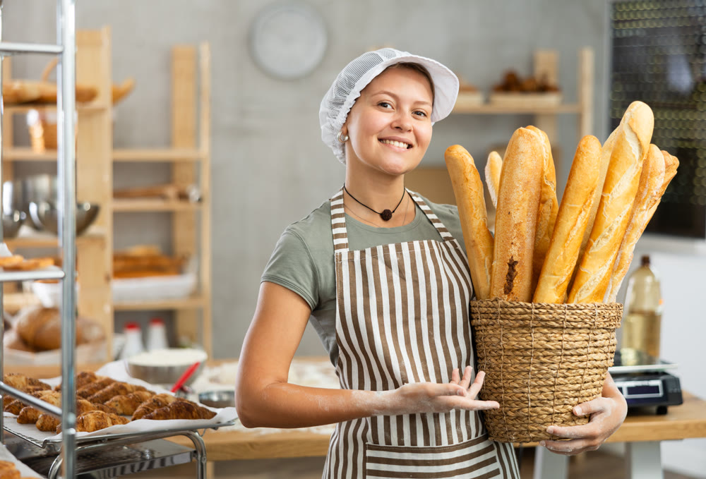 Bakery female seller stands proudly in front of fresh bread holding basket of freshly baked baguettes in kitchen