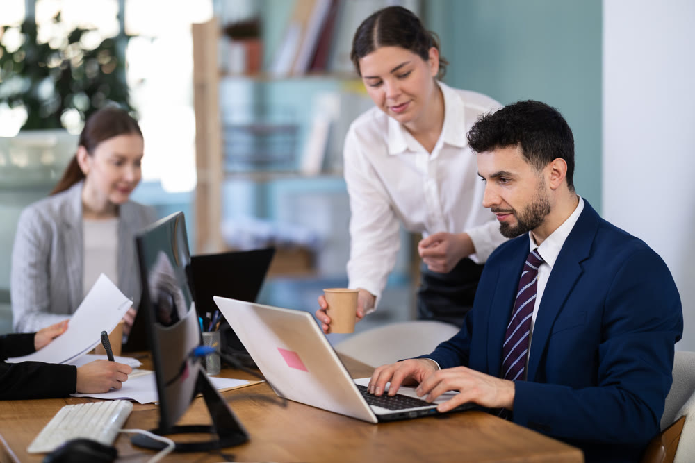 female boss helps a male employee make edits to electronic spreadsheets