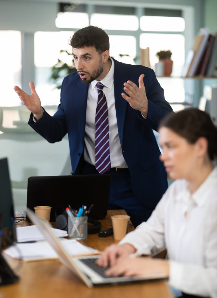 Young male manager standing and scolding colleagues during stressful corporate meeting