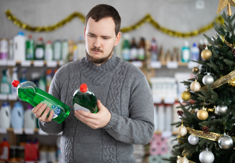 Focused young man choosing dishwashing liquid on supermarket shelf decorated for New Year holiday