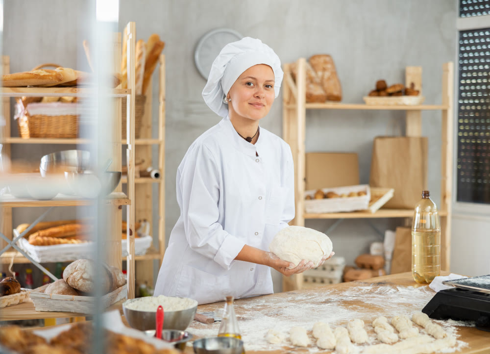 Young baker girl is standing near work table in kitchen, working with dough