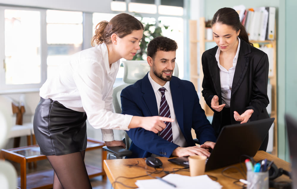 Two women help male colleague work on laptop