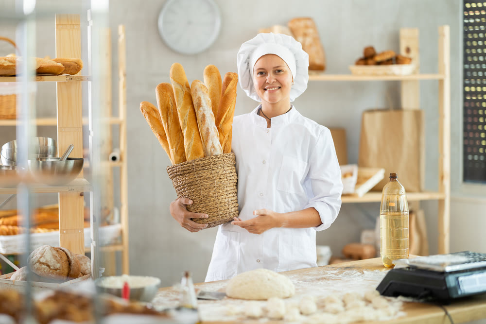 Bakery female seller stands proudly in front of fresh bread holding basket of freshly baked baguettes in kitchen