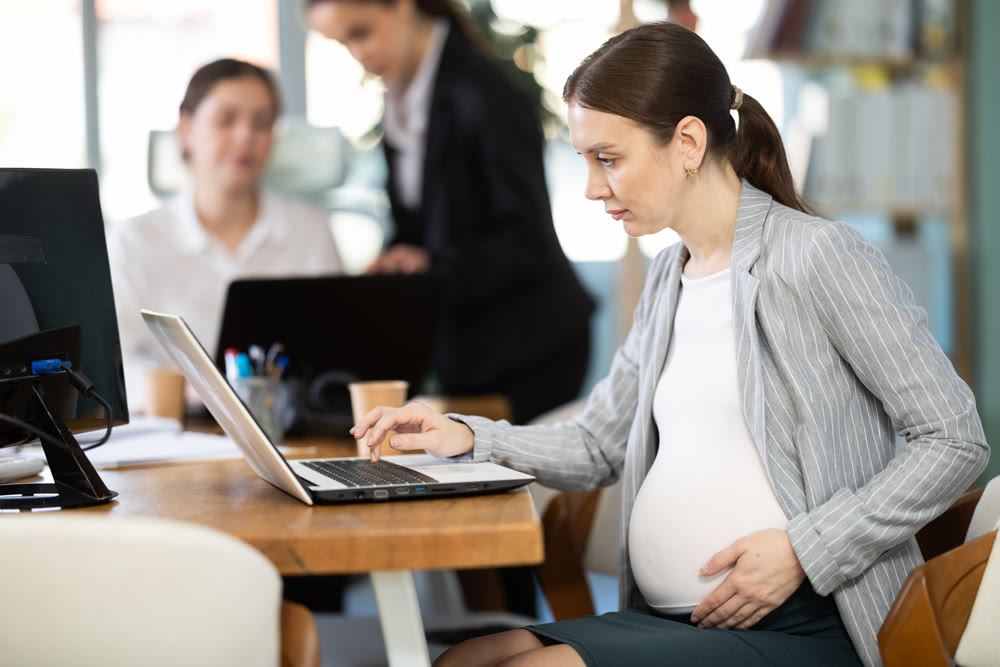 Pregnant woman is working on a laptop in an office while colleagues are discussing a project