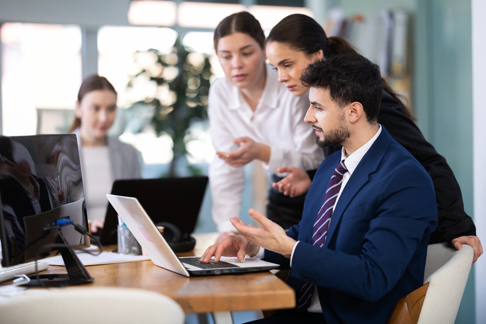 Two women help male colleague work on laptop