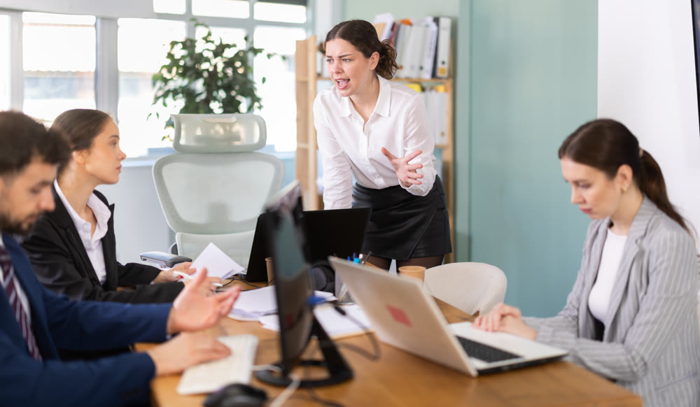 Young woman standing and scolding sad seated colleagues in office