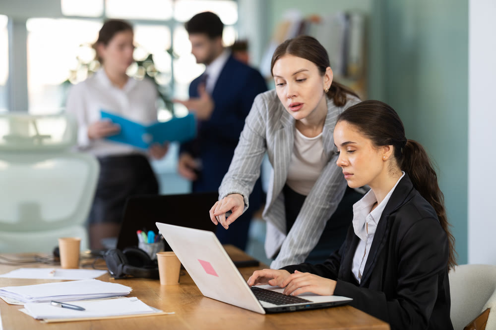 Young woman helping female colleague work on laptop