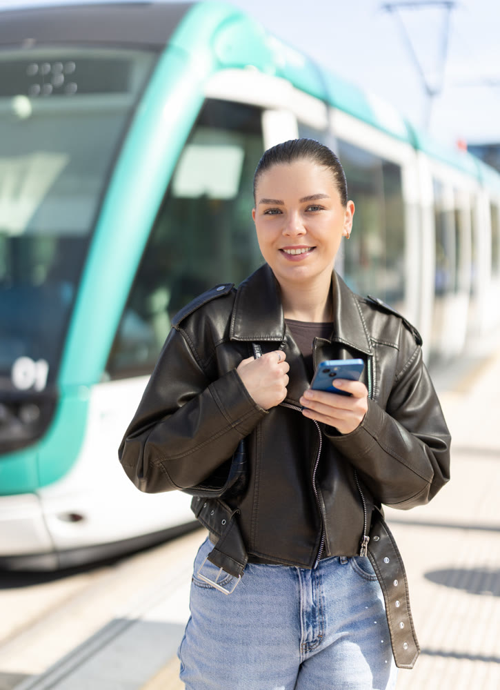 Young woman waiting for tram at stop with mobile phone