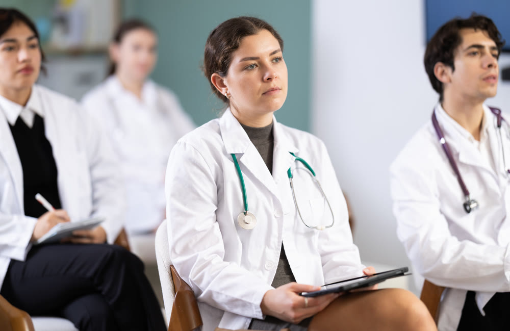 Young girl medical student dressed in labcoat sitting and listening lecture, taking notes on tablet in lecture hall
