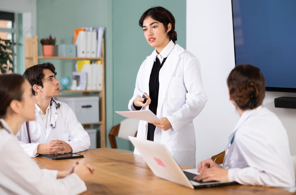 Positive young malaysian female medical student standing with papers in hands near table where sitting doctors and talking in medicals seminar in office