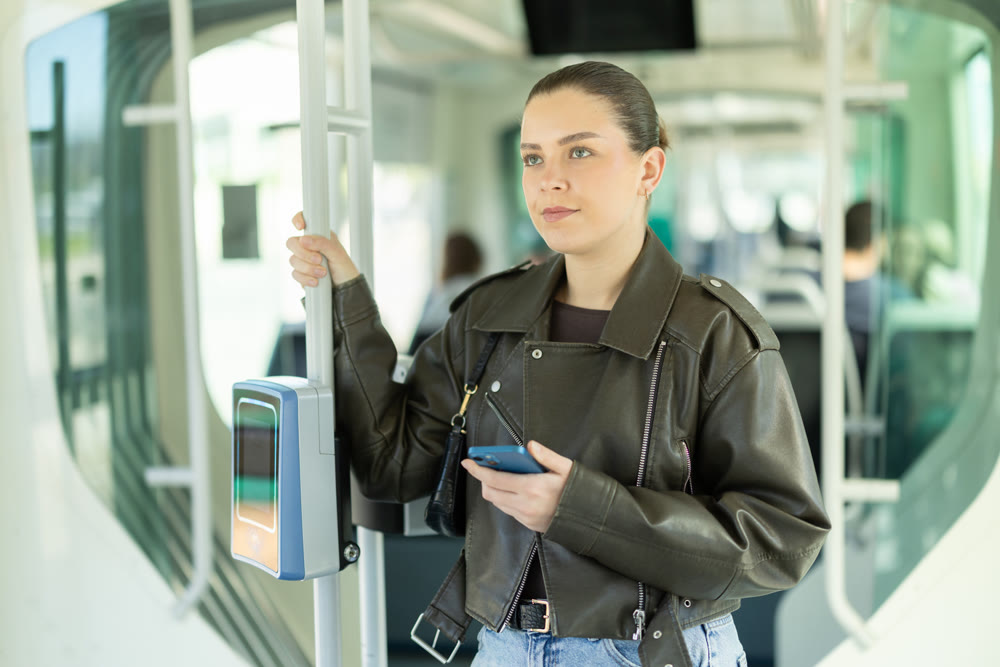 Positive woman is standing in tram wagon with phone. Girl whiles away time on trip communicating on social networks