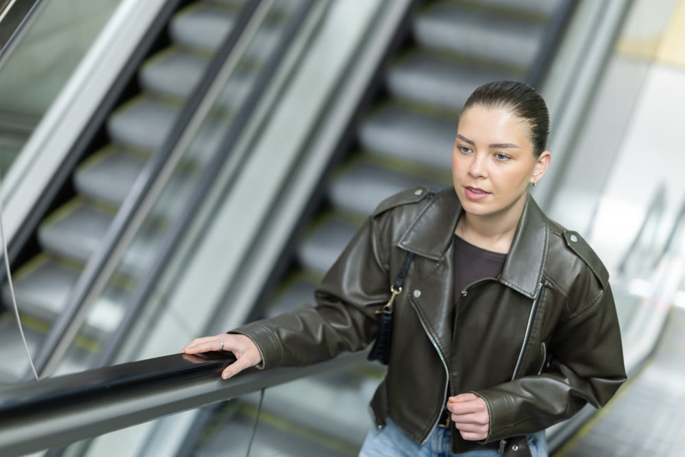 Positive young woman in black leather jacket riding up an escalator at metro station