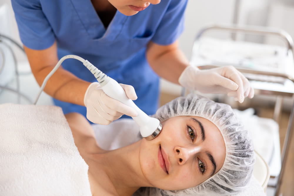 Young female cosmetologist doing ultrasound procedure to young woman
