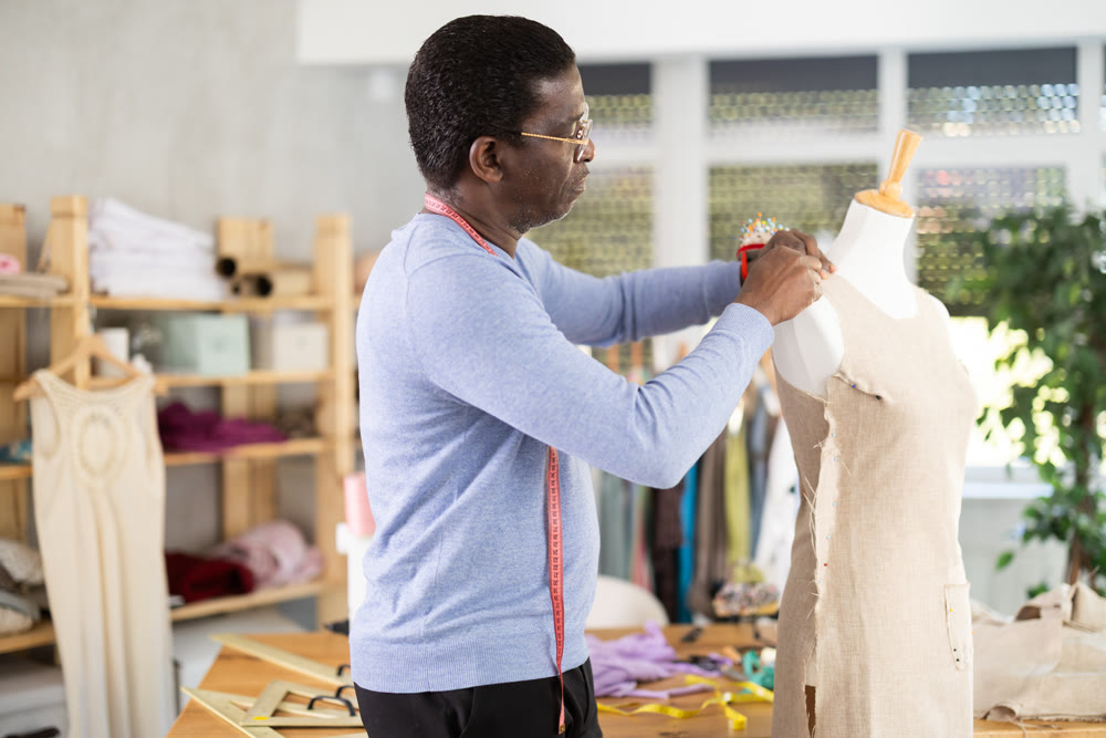 African American man tailor connects parts of dress on mannequin.
