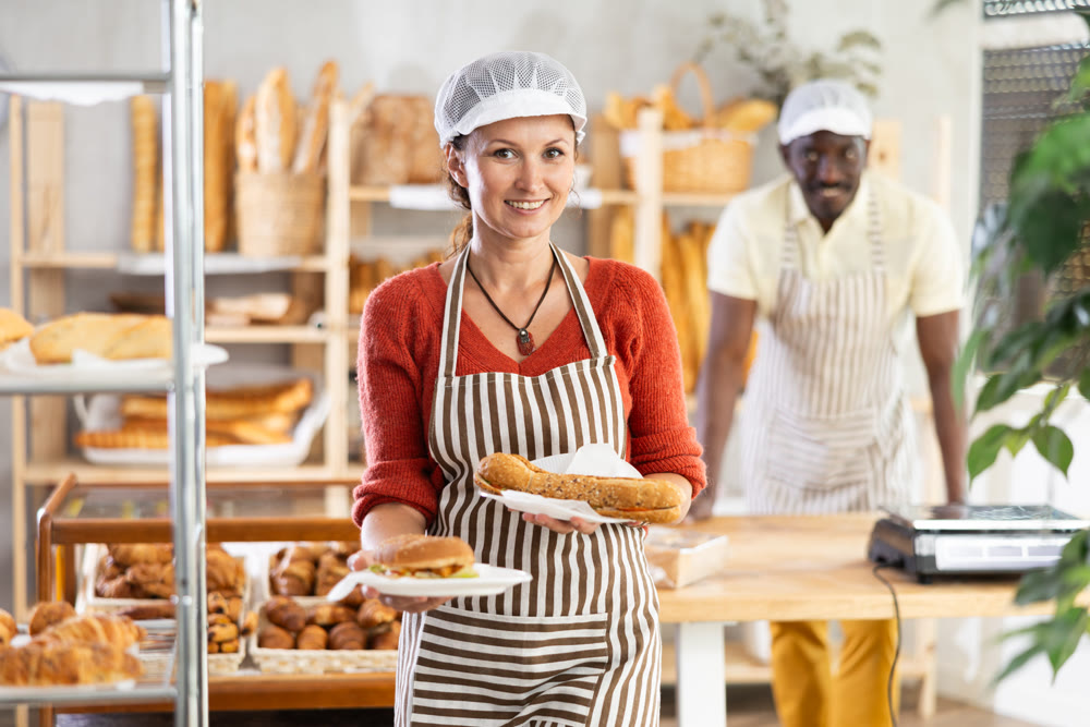 Woman entrepreneur, owner of bakery, holds out sandwich in disposable dish plate in hand