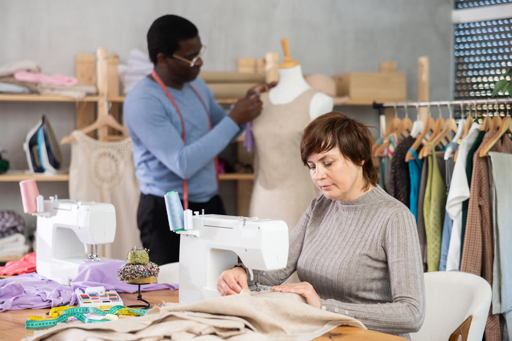 Woman tailor sews clothes, African male companion work in background with mannequin