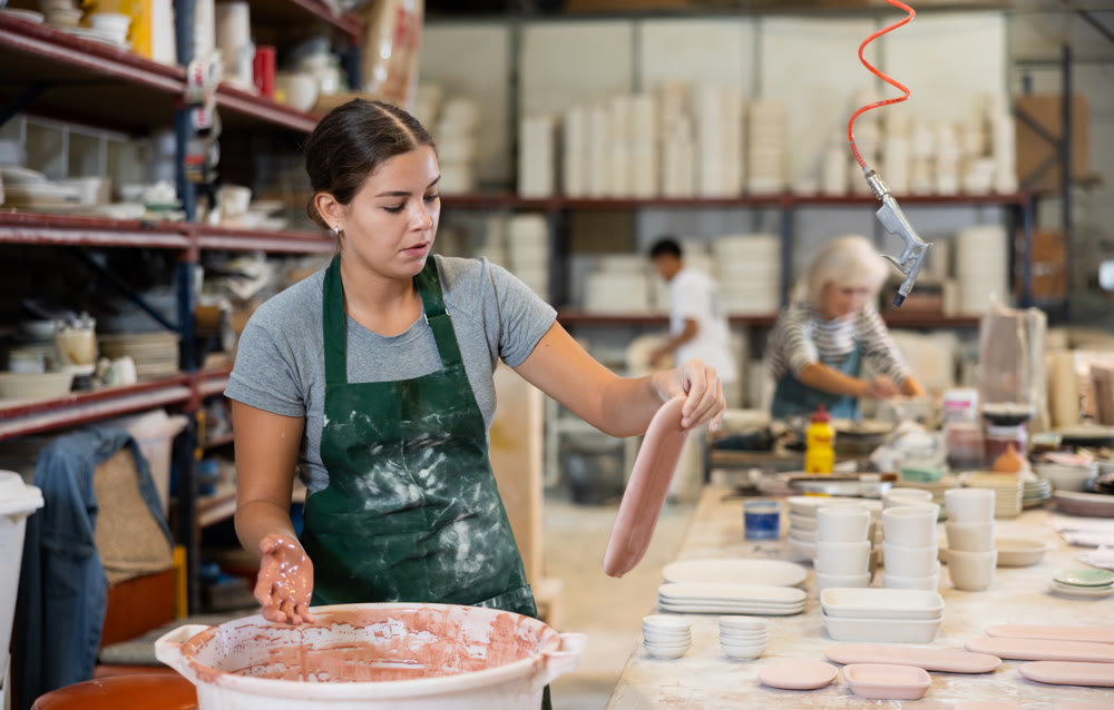 Young female worker in pottery factory lowers fired clay plates and cups into container with glaze