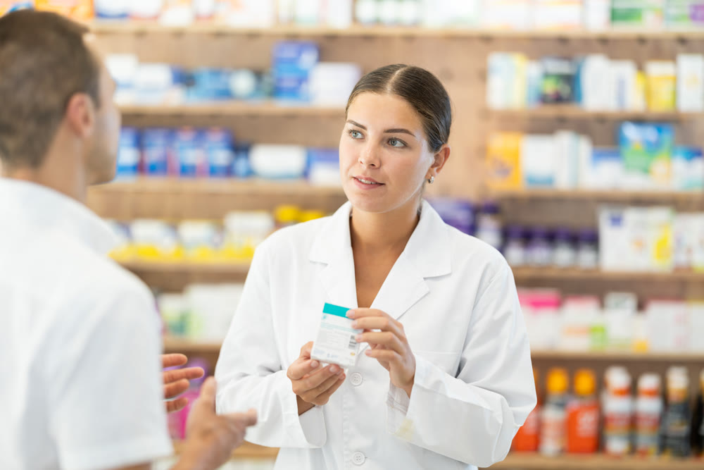 Female consultant pharmacist holds box in hands and offers man client to purchase remedy
