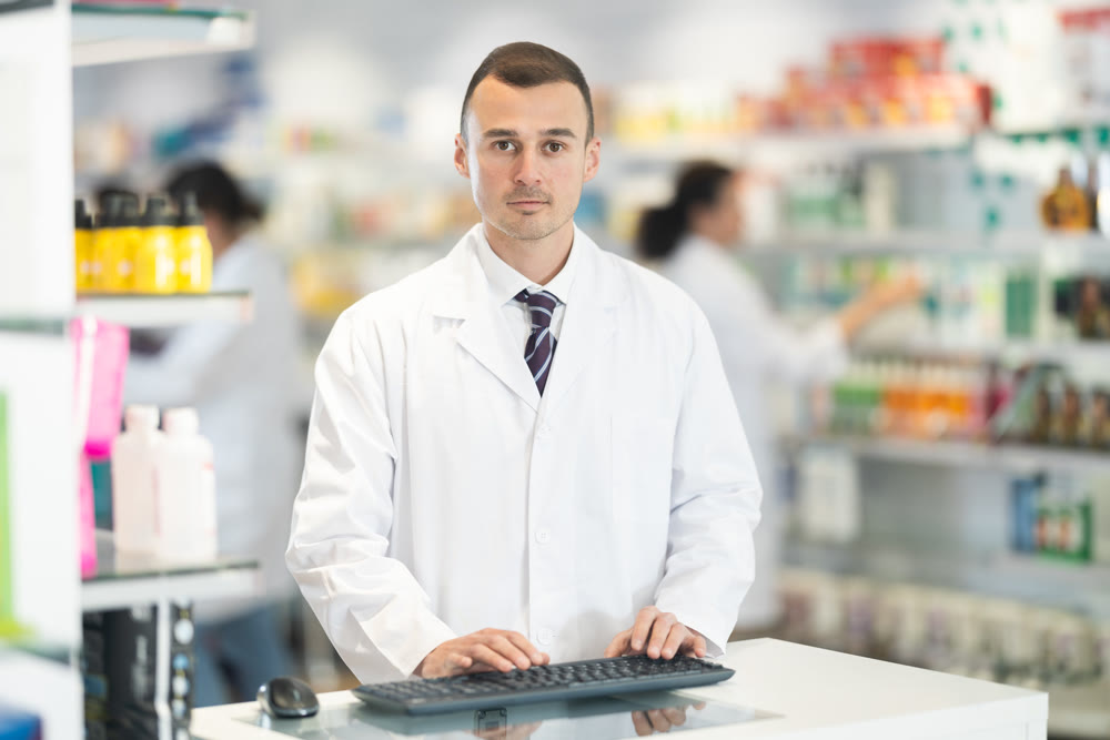 Young male pharmacist standing at the desk using keyboard in chemist's shop