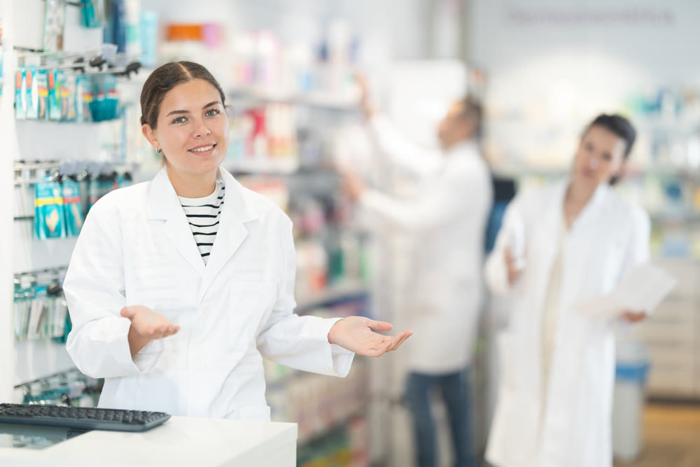 Attentive female pharmacist looking at computer in chemist shop with large assortment