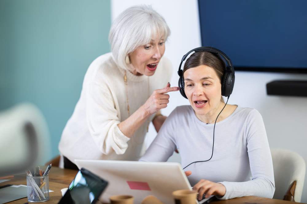 Elderly woman scolding female colleague with headphones at laptop