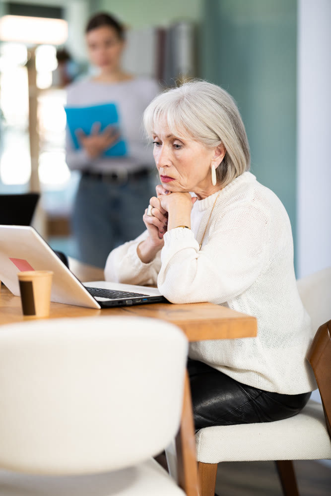 Elderly woman working on laptop adult colleague shows document