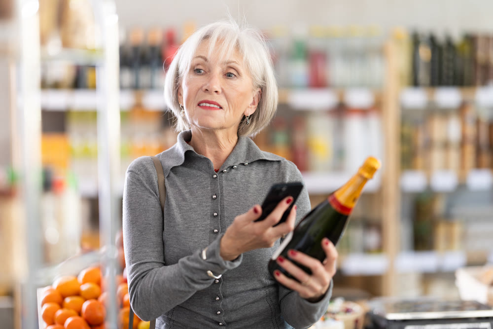 Checking expiration date of sparkling wine - elderly female shopper scans QR code on label of wine using smartphone