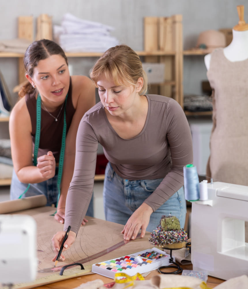 Two young female dressmakers drawing on paper in sewing workshop