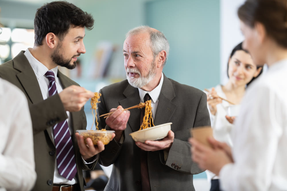 Positive two male office workers eating food together during lunch time in office of business company