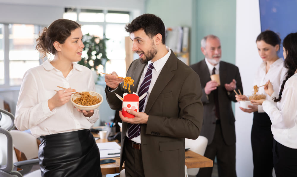 Office workers, man and girl, eating noodles during their break and chatting with each other indoor