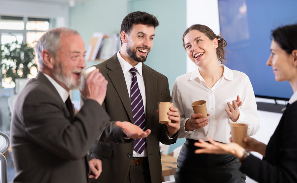 Group of positive office workers drink coffee and chat during work break in office of business company