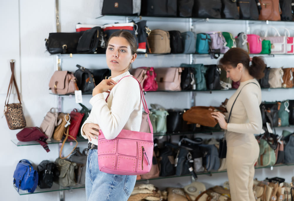 Girl stopped near accessories showcase, holds lady handbag in hands and examines product