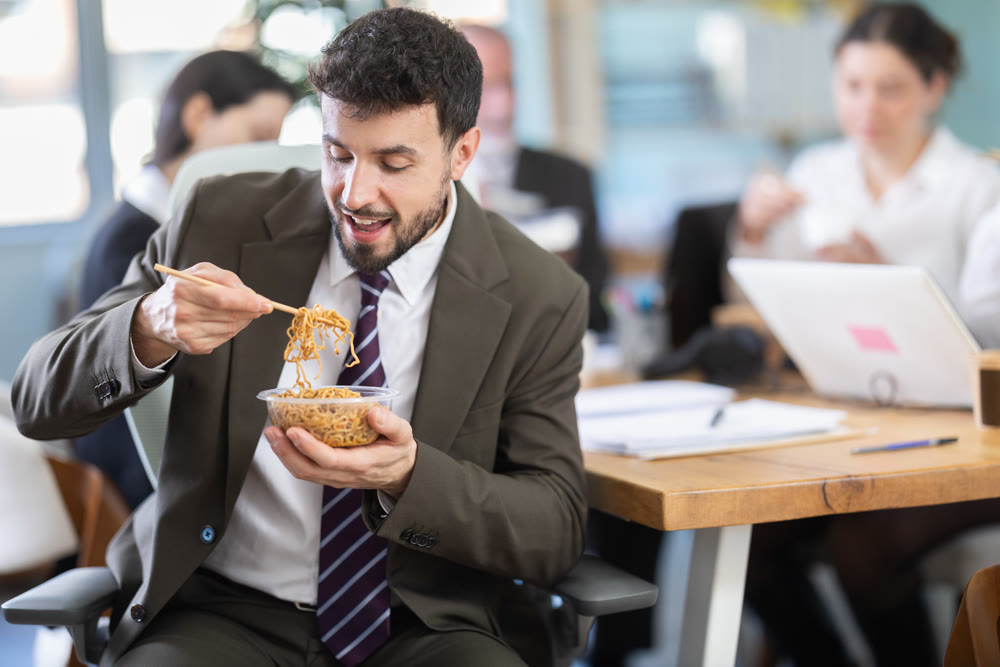 Young businessman having takeout meal during lunch break at work sitting at workplace at laptop with other colleagues in office