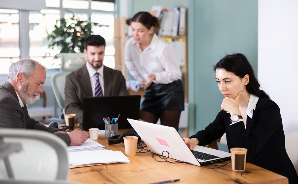 Woman sitting at desk is working on laptop computer among staff colleagues.
