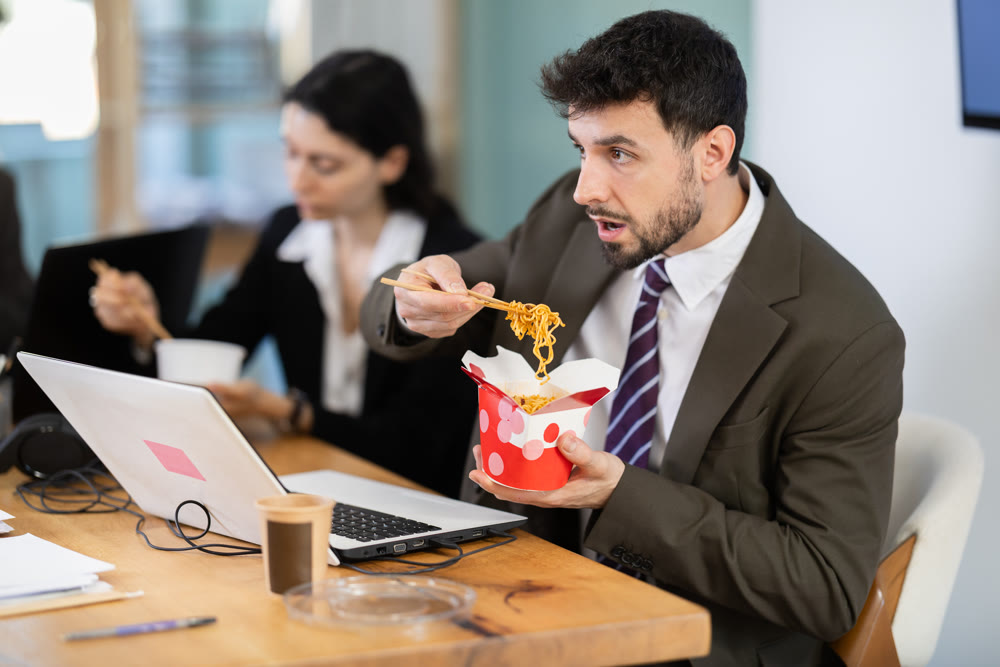 Young businessman having takeout meal during lunch break at work sitting at workplace at laptop with other colleagues in office