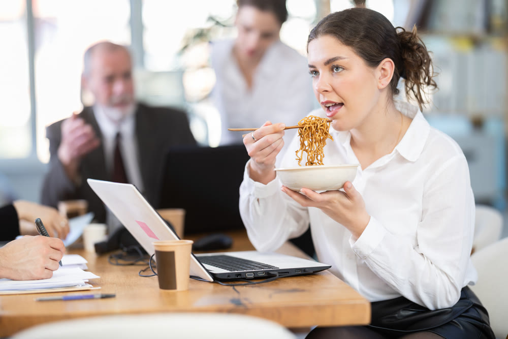 Positive young female office worker eating food at workplace, while other co-workers unhappy and continue to work in bright office of business company