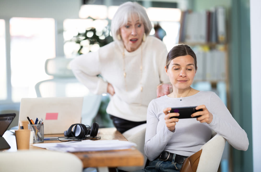 Elderly woman scolding adult female colleague for sitting with mobile phone