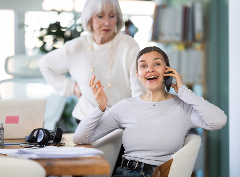 Elderly woman judging female colleague talking on mobile phone
