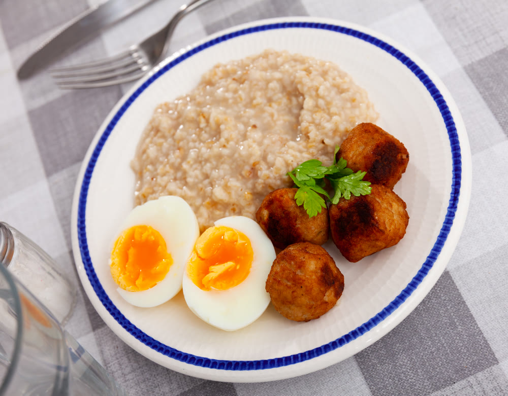 Homemade breakfast is served on plate - small portion of oatmeal, stewed meatballs and boiled egg
