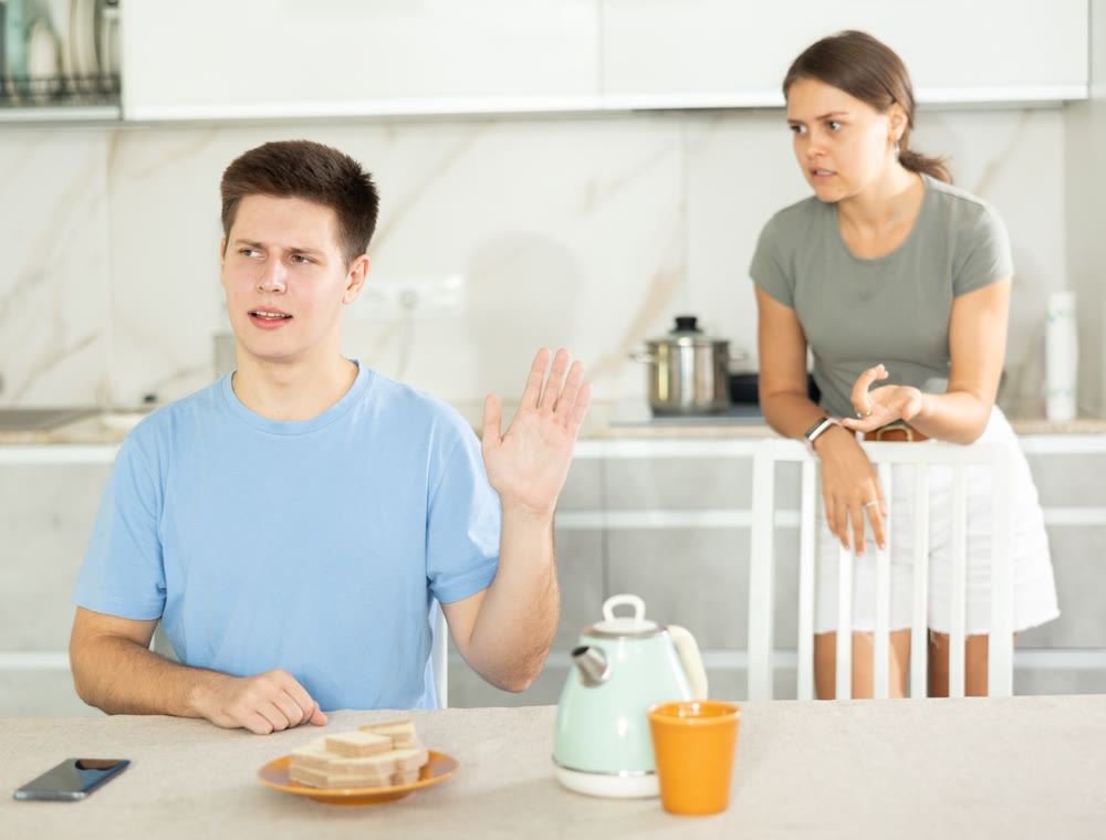 Young guy sitting at the table while his girlfriend is quarreling with him standing behind