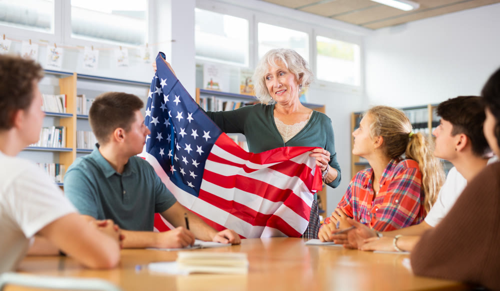 Mature female teacher shows scholchildren the national flag of USA