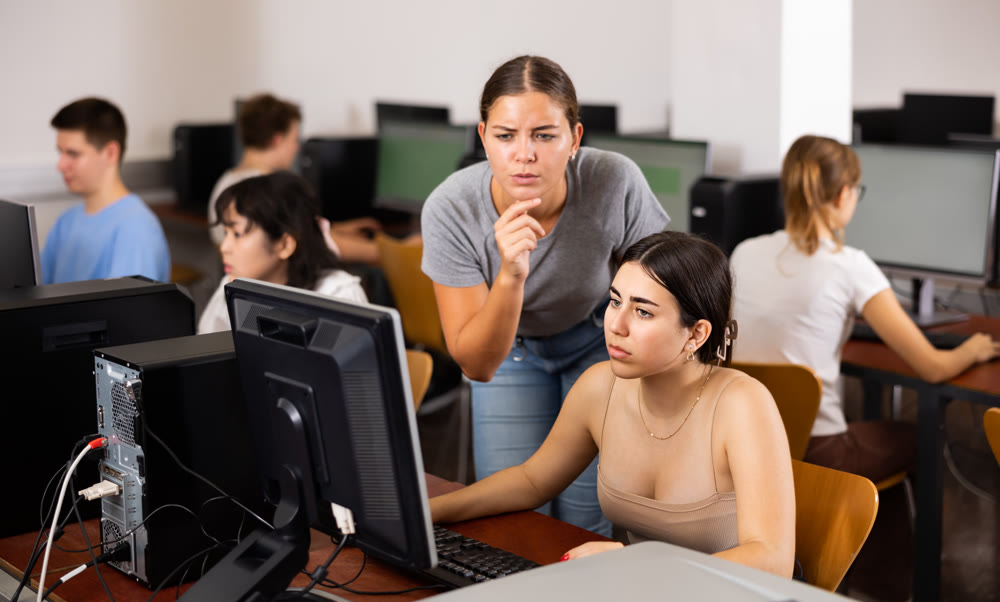 Focused young woman student working with her female friend in computer class, using pc in library