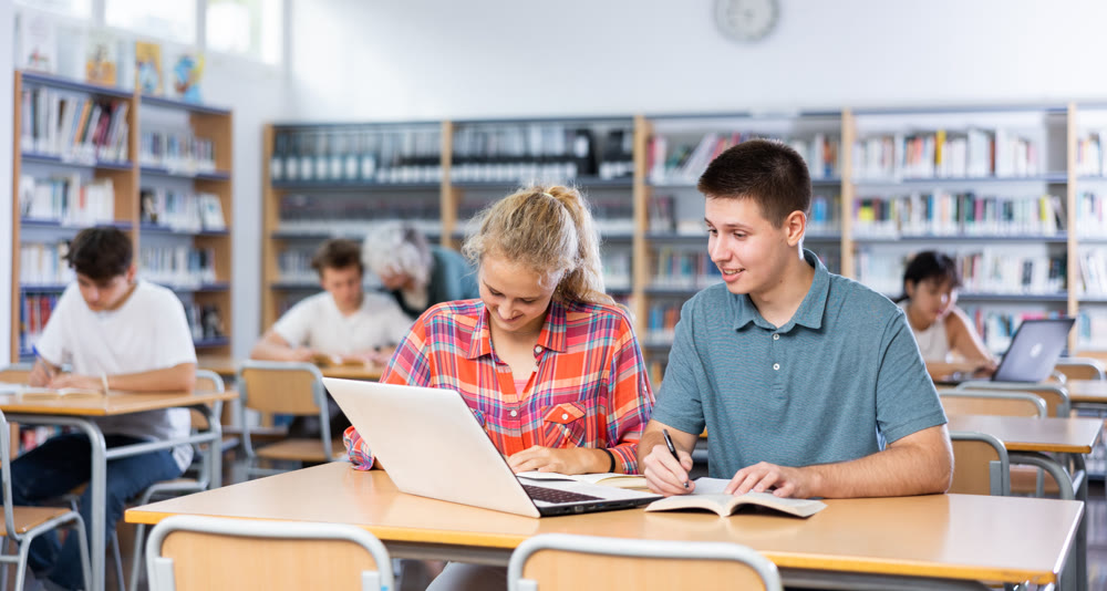 Teenage students help each other study at laptop while sitting in school library