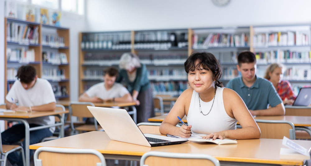 Concentrated asian teenager female schoolgirl sitting at library desk, doing homework with internet