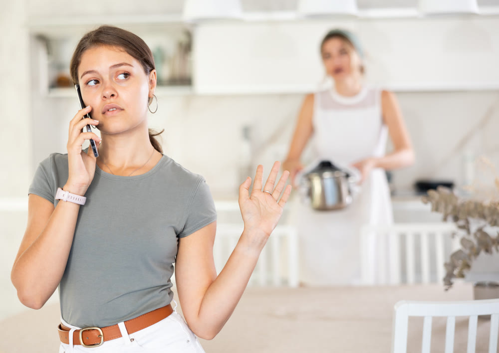 Upset daughter is sitting at table. Mom yells at her daughter in background